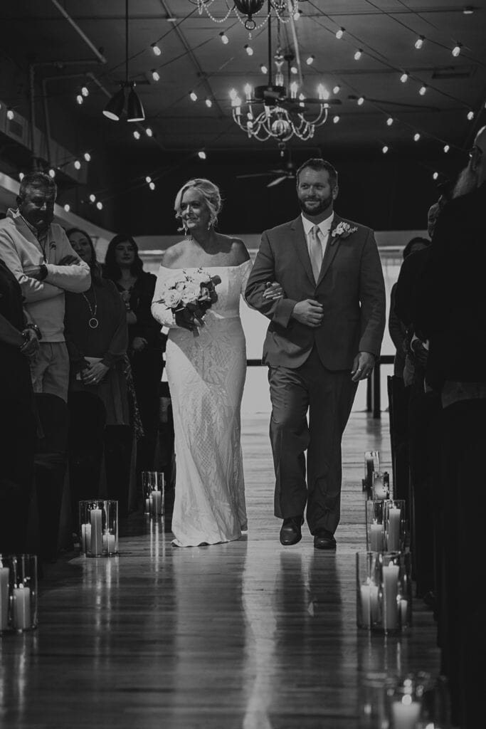 A black and white image of a son walking his mom down the aisle of her wedding. Candles are lighting the floor of the aisle.