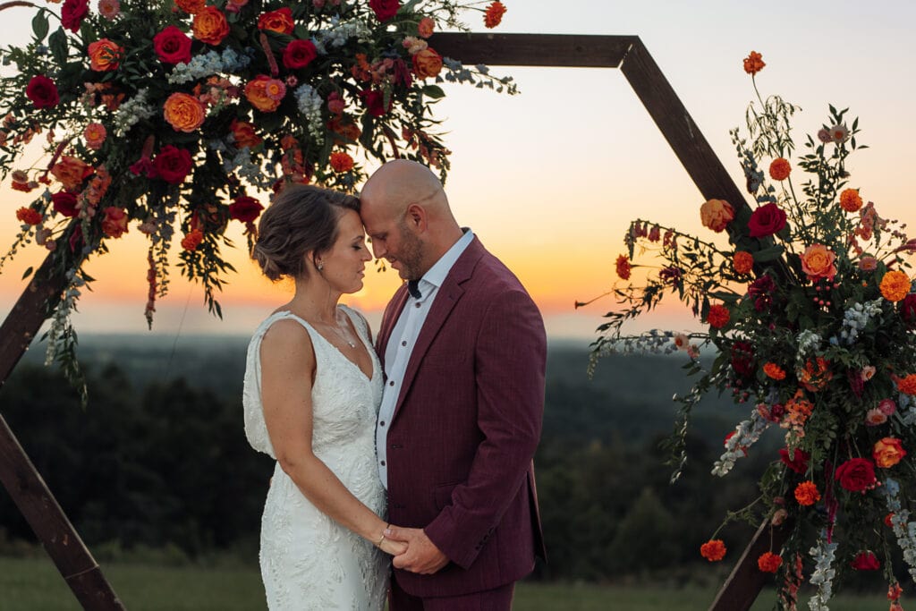 A Bride & Groom stand facing each other, holding hands, and touching foreheads. It is sunset and an octagon arch with flowers is behind them.