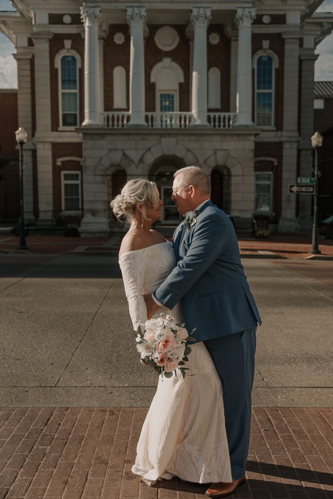 Groom leans into Bride as she holds her bouquet at her side. A historic courthouse is in the background.