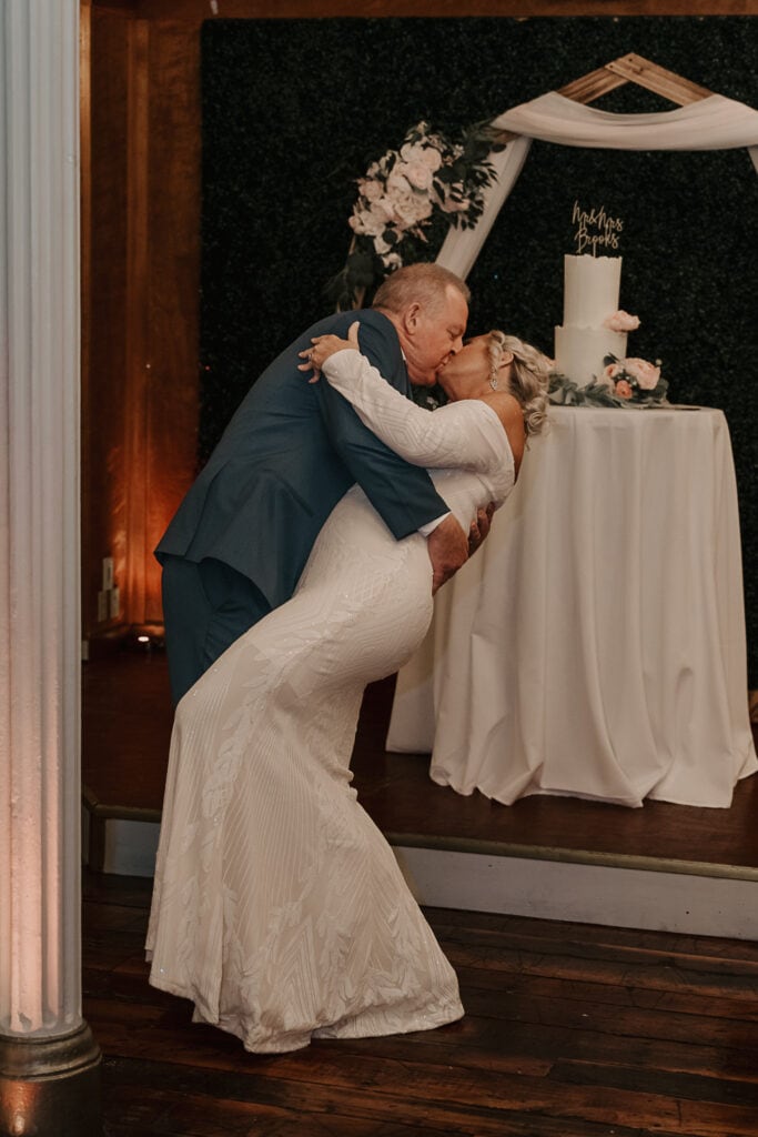 Man dip kisses bride during their first dance. Their wedding cake is in the background.