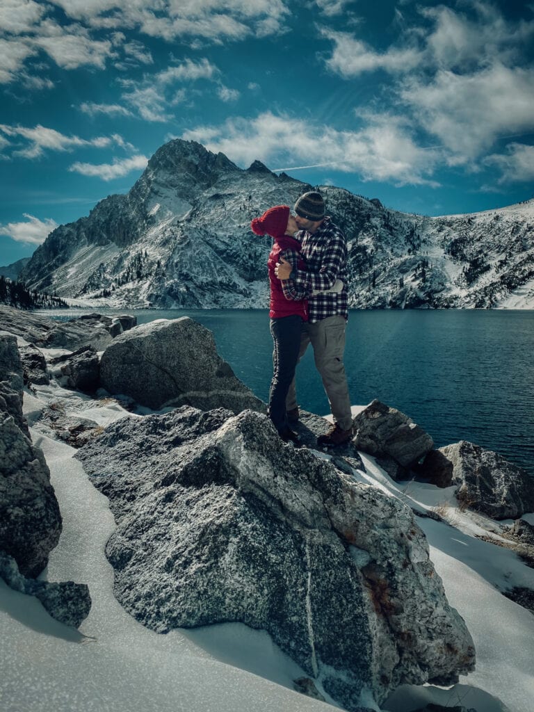 Korey and Jeff kiss on top of a rock in front of Stanley Lake to celebrate their hike to the top of the mountain in the snow.