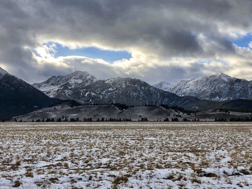 View of a snowy field and the Sawtooth mountains as the blue sky peaks through the clouds.