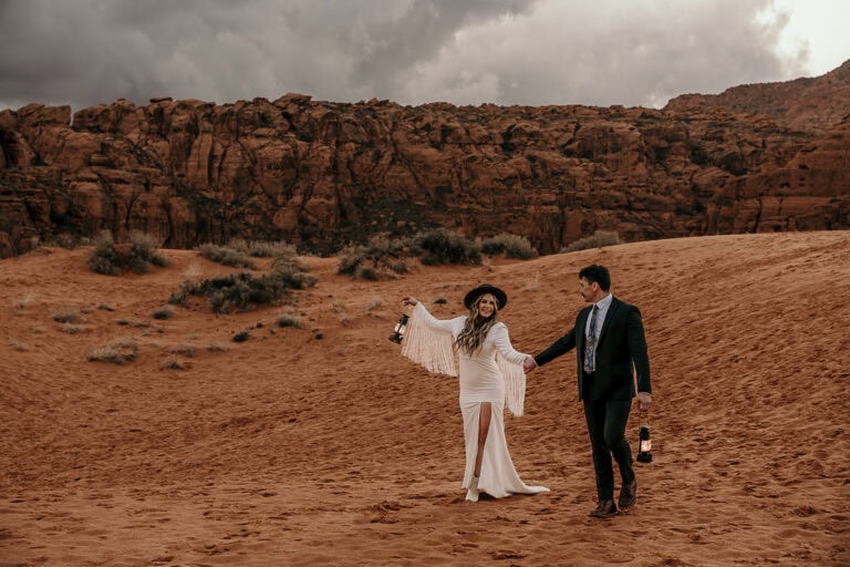 A couple in wedding attire walks across sand dunes while swinging lanterns.