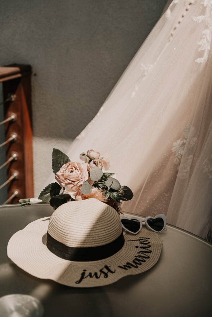 A Bride's sunhat and heart shaped glasses set on a table with her wedding dress hanging behind them.