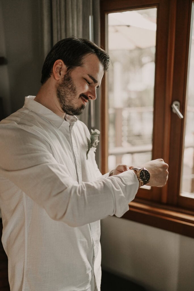 Groom adjusts his cuffs in front of a window.