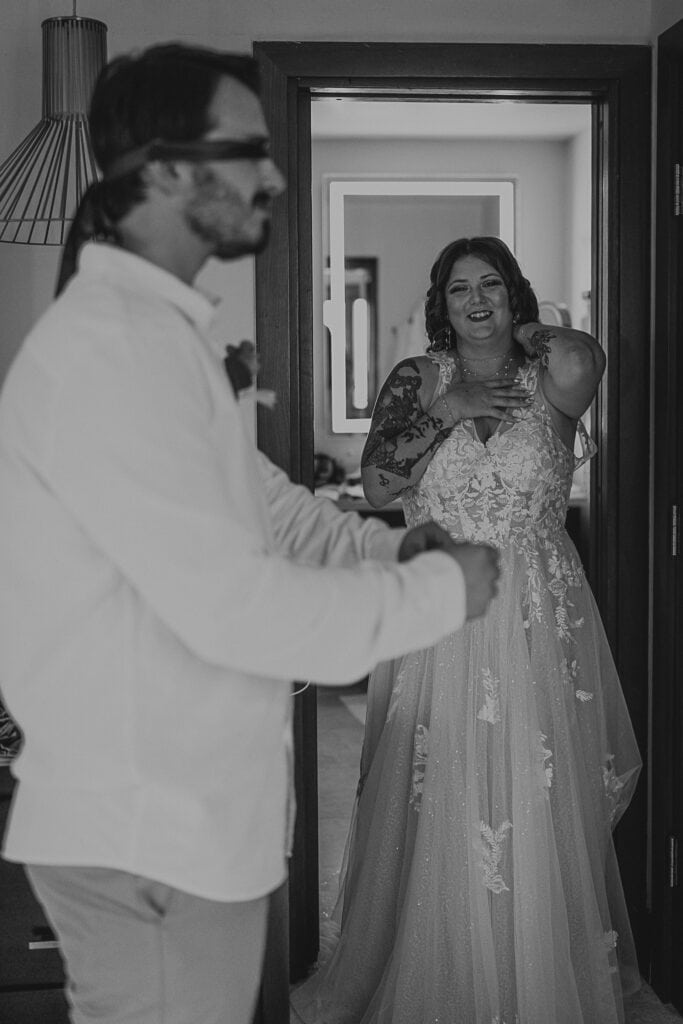 A bride in her wedding gown smiles at the groom who is wearing a blindfold so he can't see her.