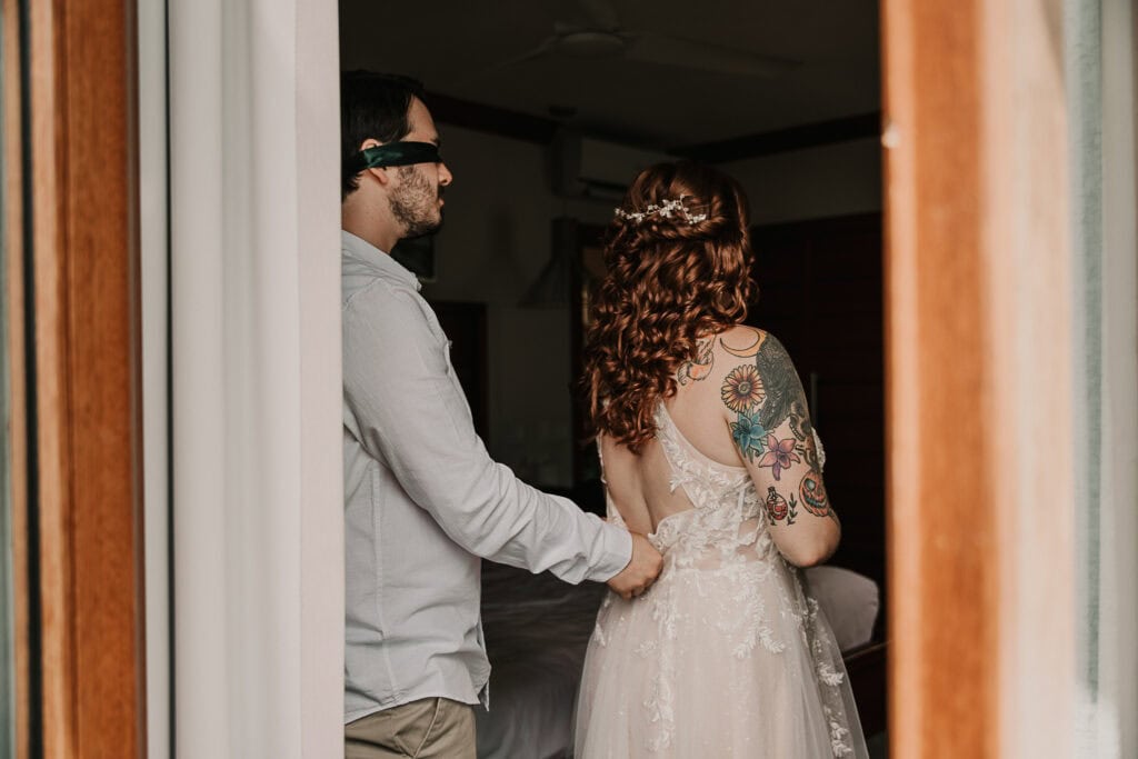 A blindfolded groom helps the bride zip her dress.