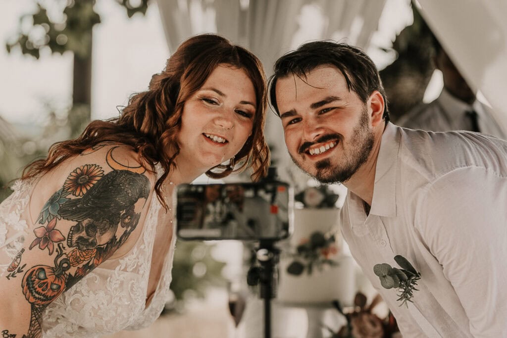 A couple smiles at their family and friends who are watching their wedding livestream on a phone.