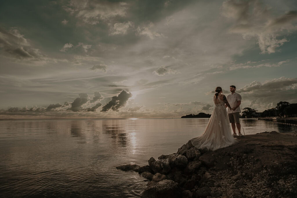 A bride and groom stand on rocks as the sunsets. The ocean is behind them.