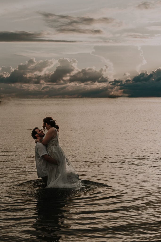 Groom lifts the bride up as they play in the ocean in their wedding attire.