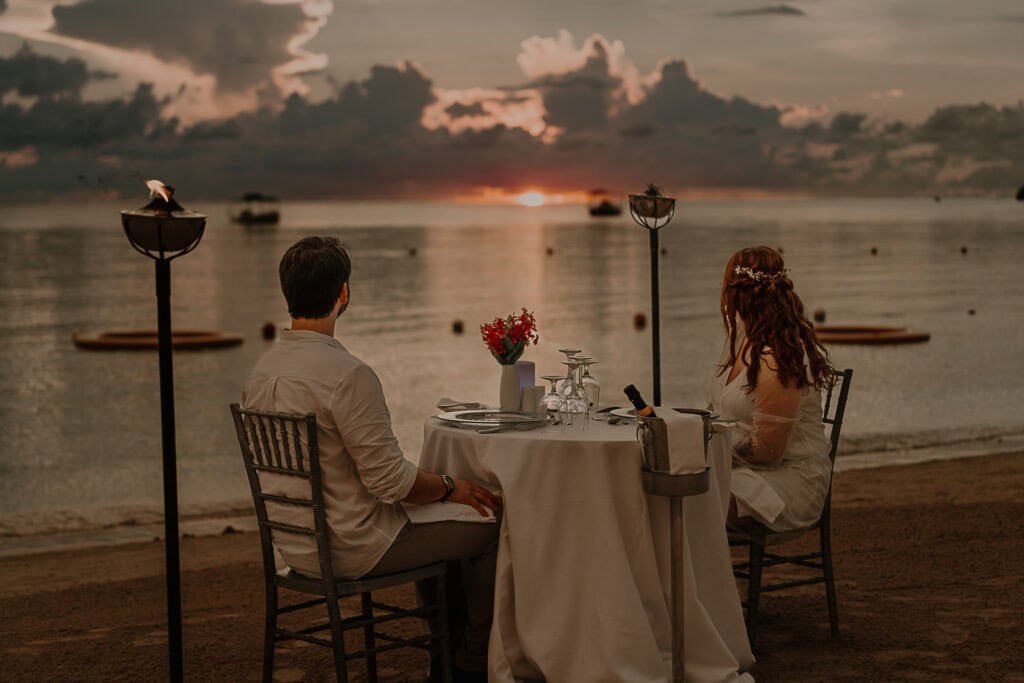 A bride and groom watch the sunset from their candlelight dinner table on the beach.