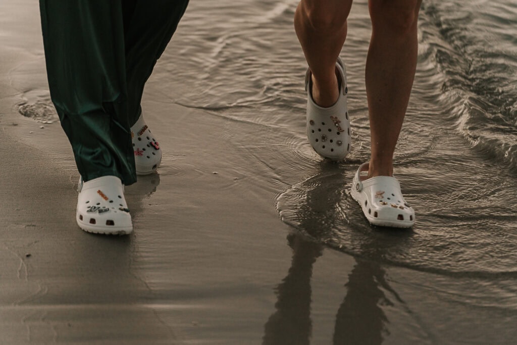 A couple walks along the beach in white wedding crocs.