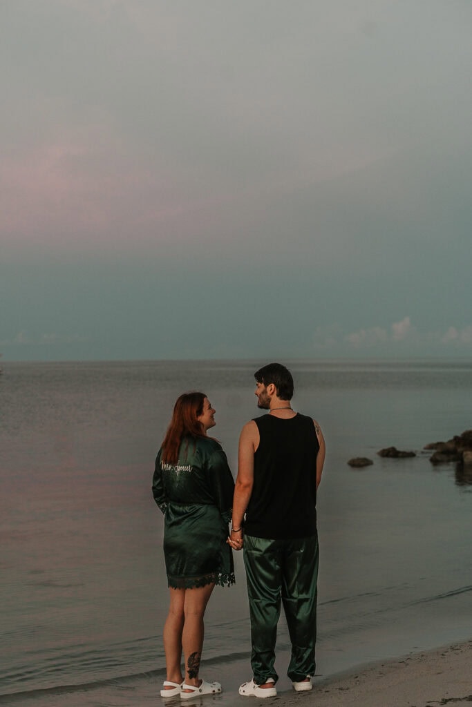 A couple holds hands while standing on the beach at sunrise.