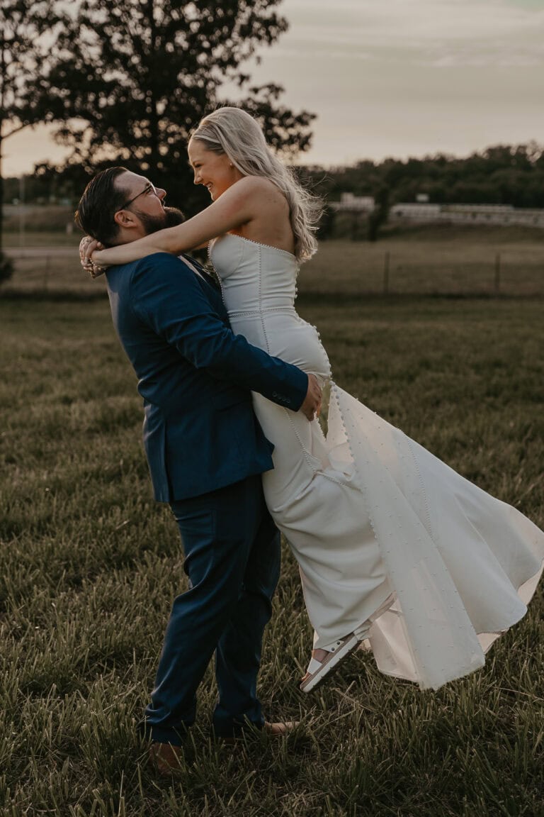 Groom lifts the bride up during sunset, as they look happily at each other.