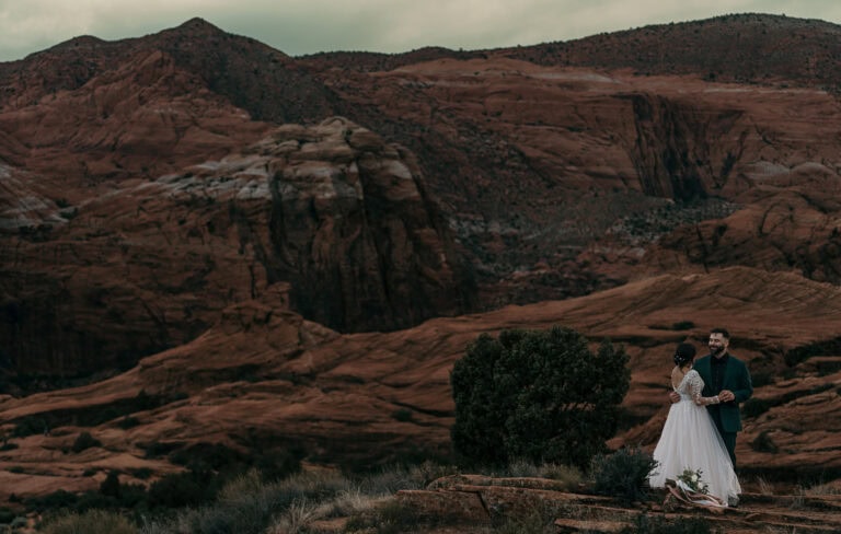 Bride and groom embrace while standing on red rocks during their wedding.