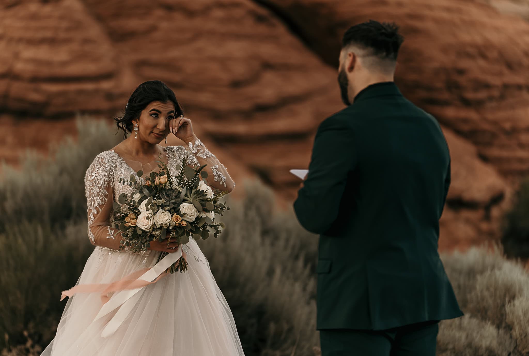 Bride wipes away a tear as the Groom says his vows in front of red rocks.