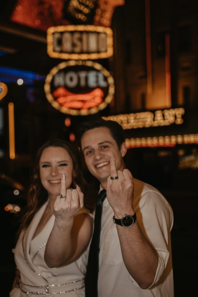 Couple flashes wedding ring fingers at camera in front of Las Vegas sign.