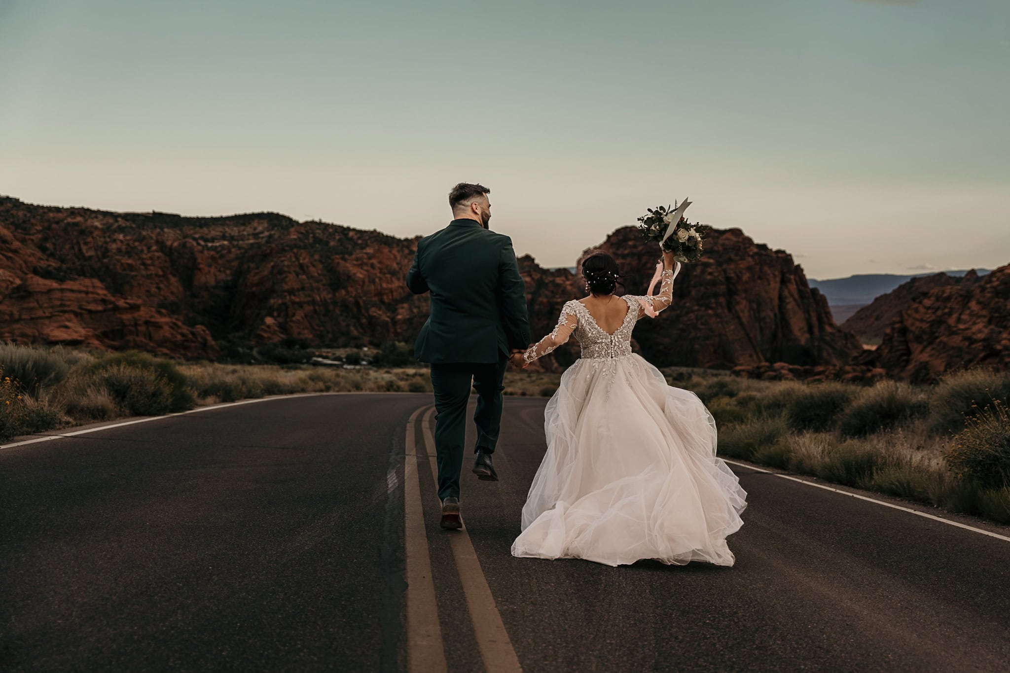 A happy couples runs down the middle of the road in wedding attire.