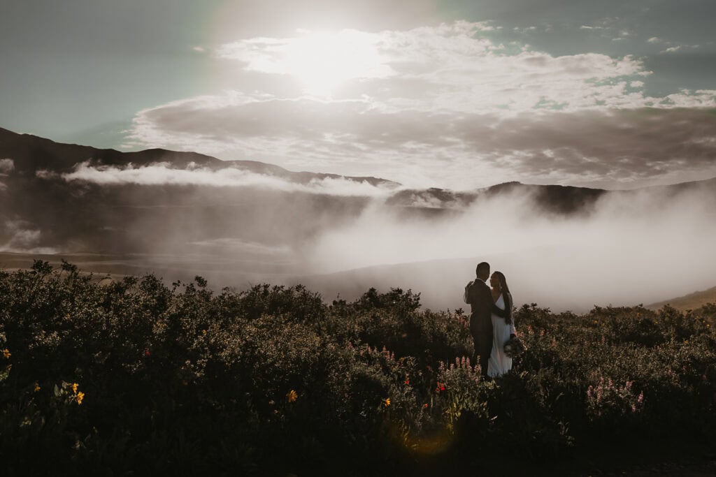 A couple in wedding attire stand on top of a mountain as fog rolls in at sunrise.