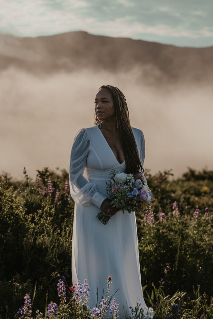 A bride stands in a wildflower field at sunrise with fog behind her.