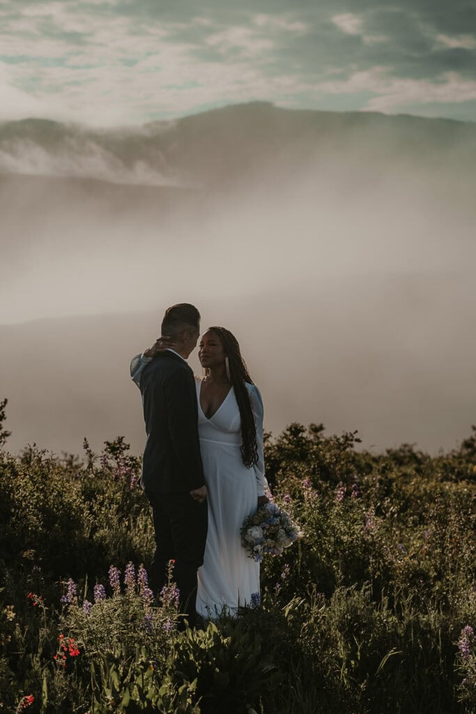 A couple faces each other on top of a foggy mountain. They're in wedding attire.