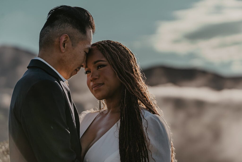 A bride and groom stand forehead to forehead as fog rolls behind them on the mountaintop.