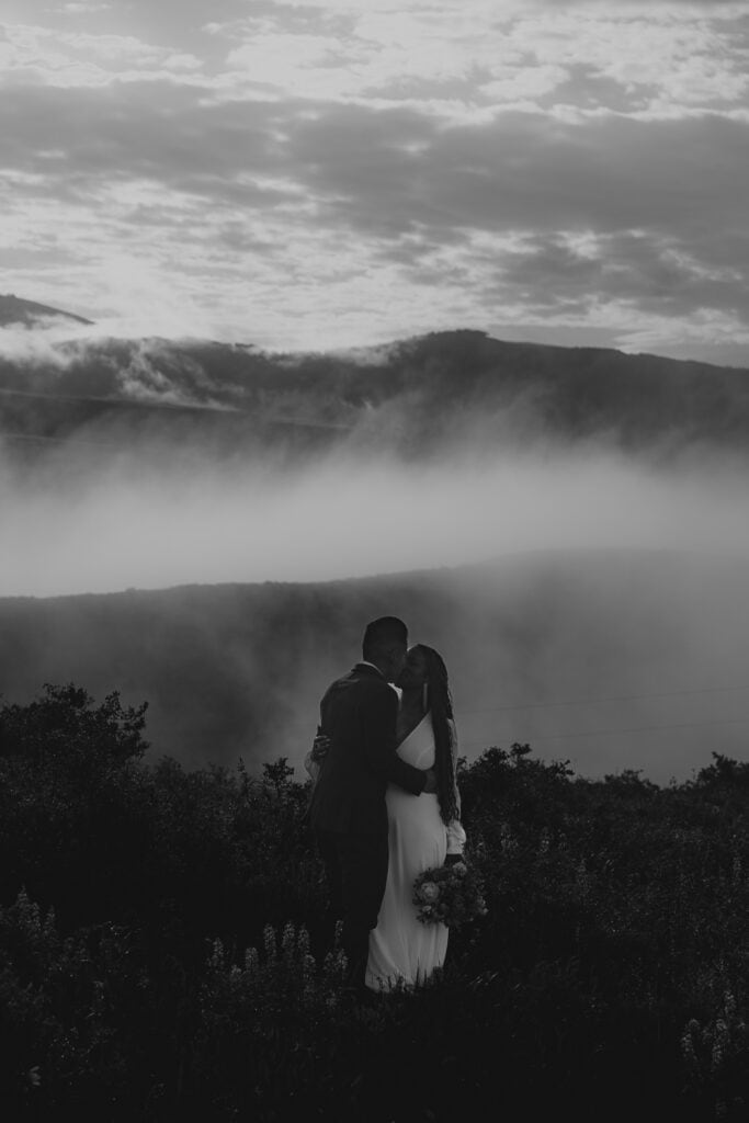 A black and white image of a couple in wedding attire on a foggy mountaintop.