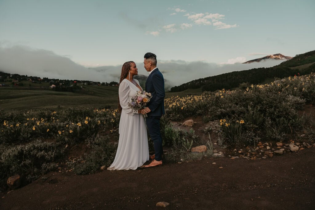 Marissa & Janiel stand on their wedding day, in a field of wildflowers with mountains in the distance.