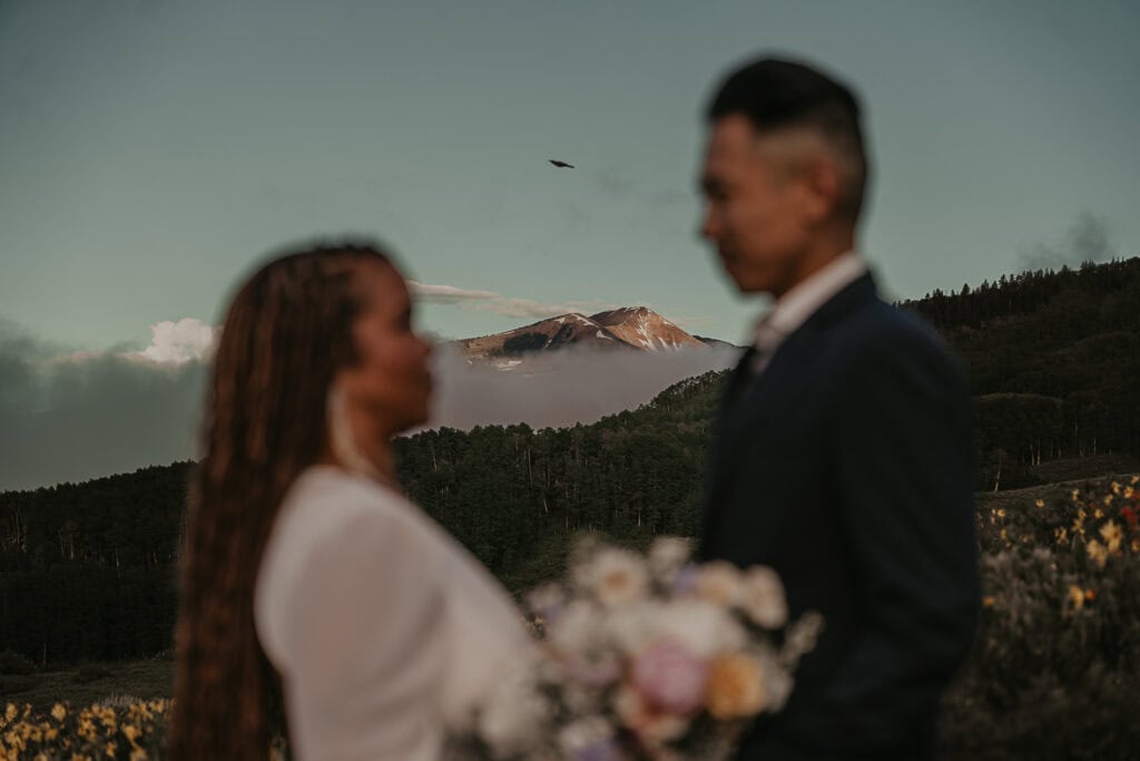 A could in wedding attire face each other. They're out of focus and the focus is on a mountain in the distance behind them.