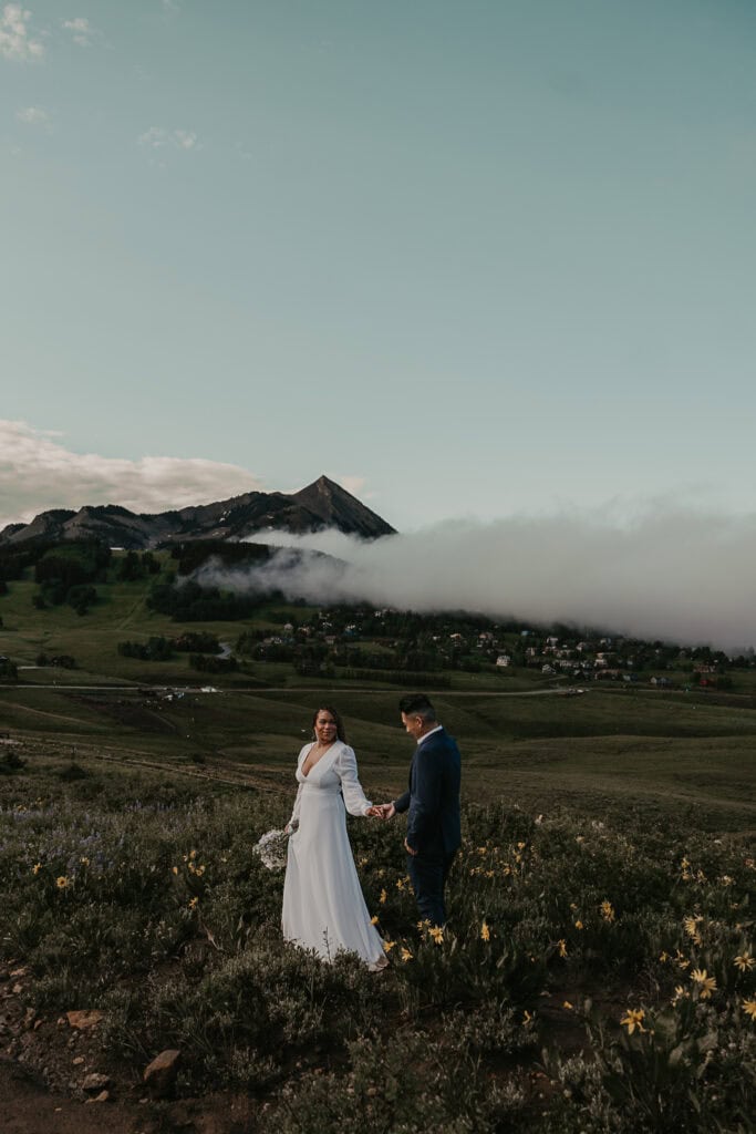 Marissa & Janiel walk hand in hand on a path of wildflowers. Foggy mountains are in the background.