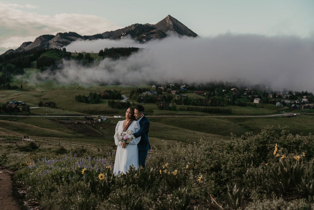 A groom hugs the bride from behind. Mountains and fog are in the distance.