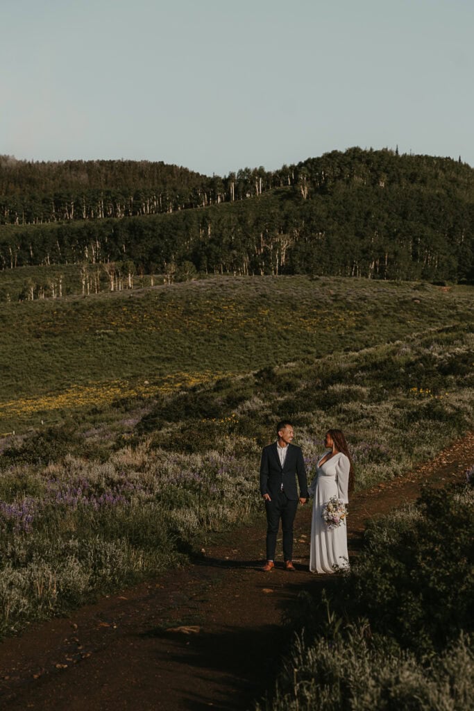 A bride and groom stand on a dirt path, laughing together. Wildflowers surround them.