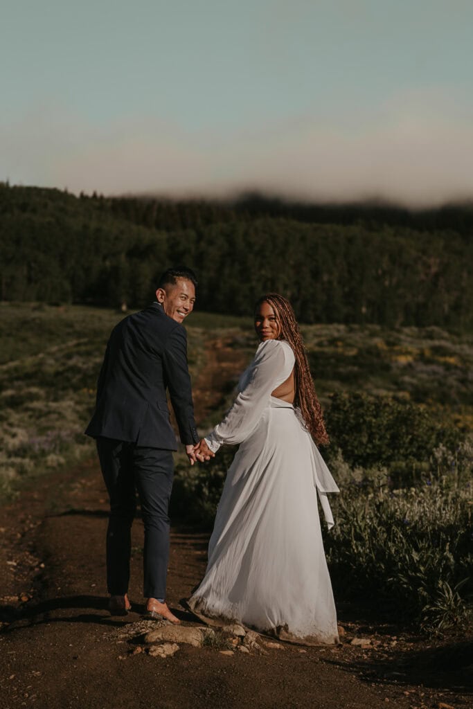 A couple looks back at the camera as they walk away while holding hands. Mountains are in the background.