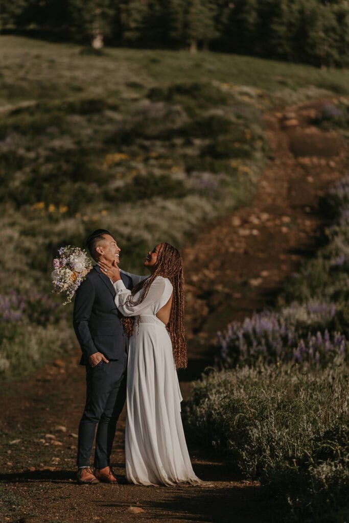 A couple laughs hysterically during their elopement. They're outside surrounded by wildflowers.