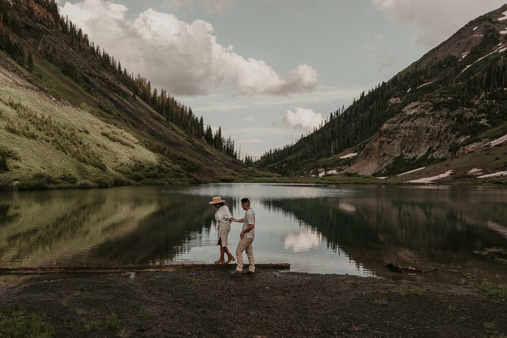 A couple holds hands while walking in front of an alpine lake. Mountains are in the background.