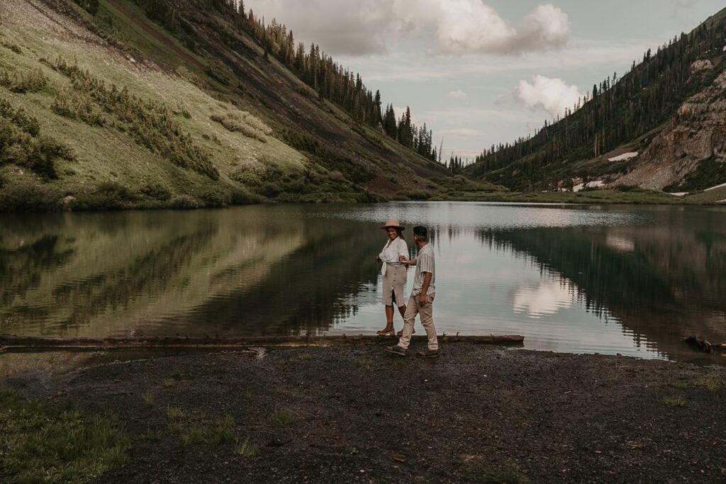 A couple walks in front of mountains and a lake while holding hands.