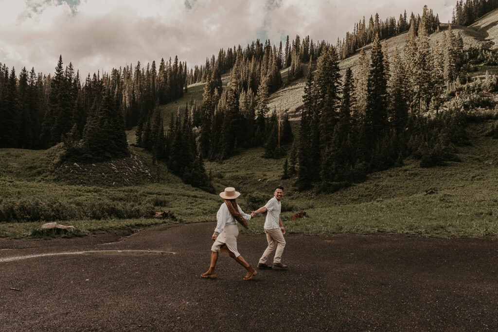 A couple runs playfully in front of mountains and pine trees.