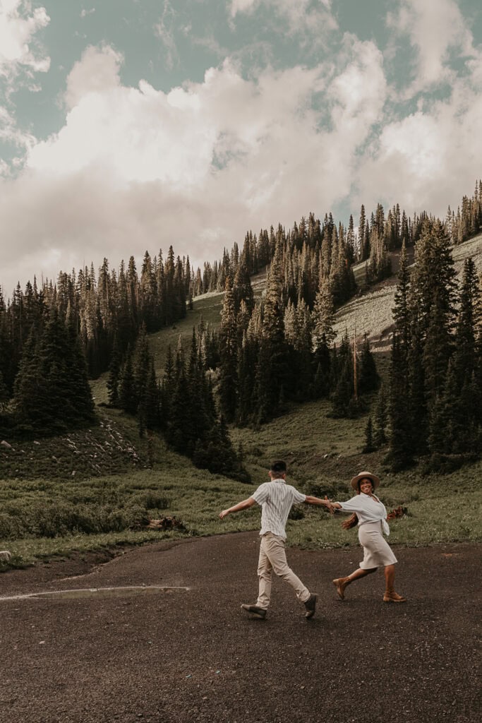 A couple playfully runs towards each other and misses grabbing each others hands. Mountains and pines are in the background.