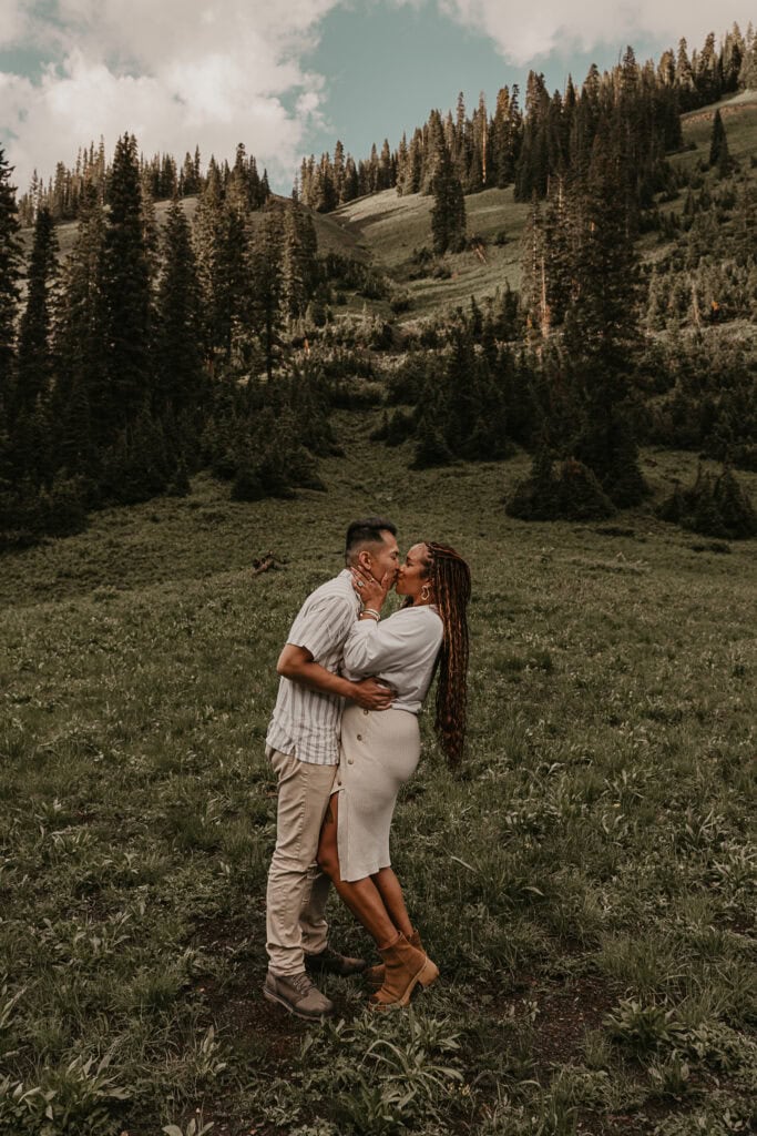 A couple kisses while standing in a field. Mountains and pine trees are in the background.