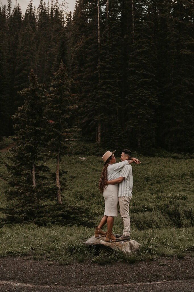 A couple stands on top of a rock with their arms around each other. Pine trees are in the background.