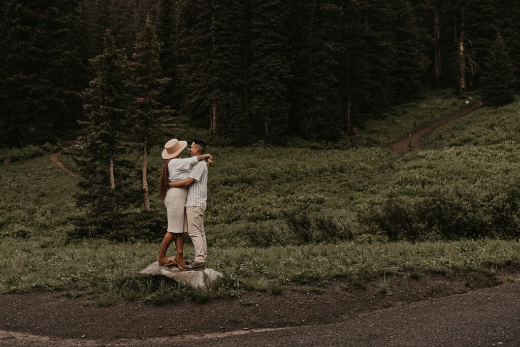 A couple stands on a large rock in the forest, embracing.