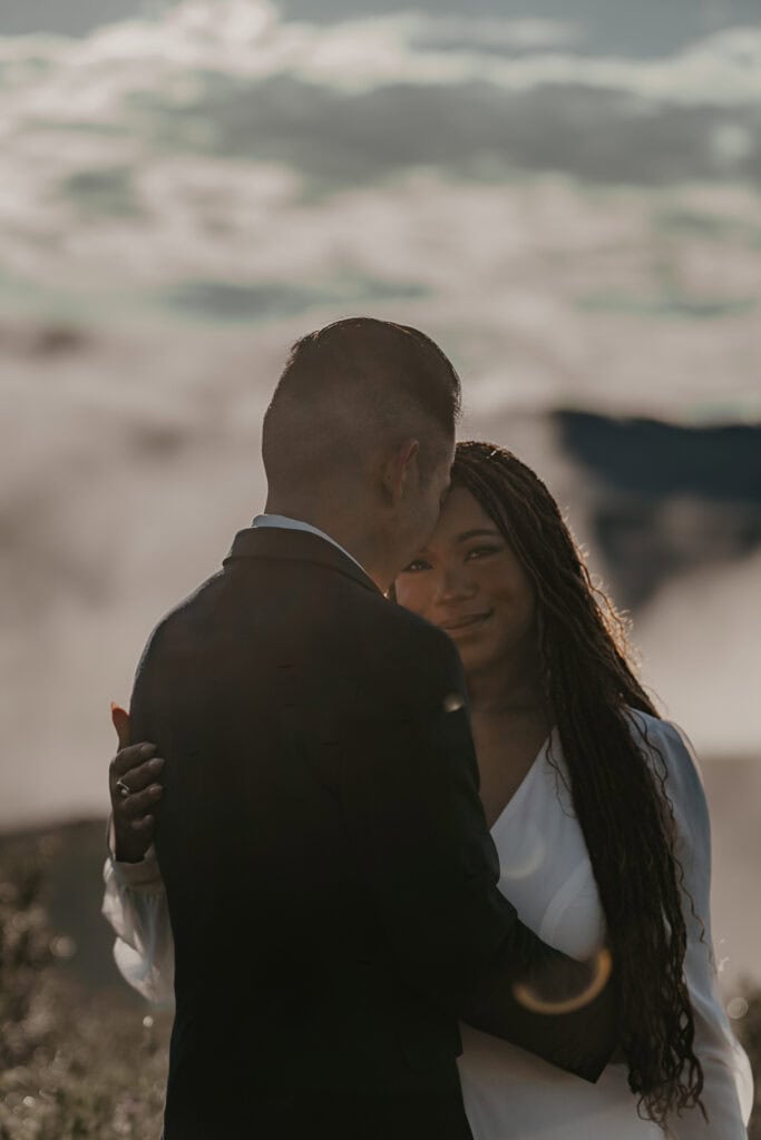 A couple in wedding attire embrace on a mountain top as fog surrounds them.