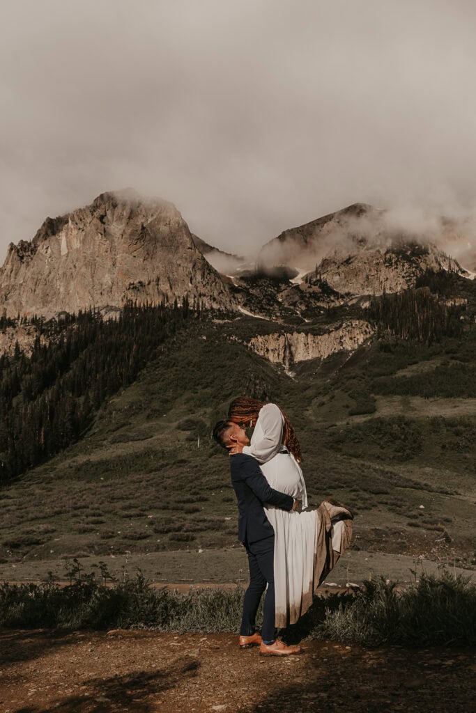 A groom lifts the bride up as they kiss during their elopement. Fog covered mountains are behind them.