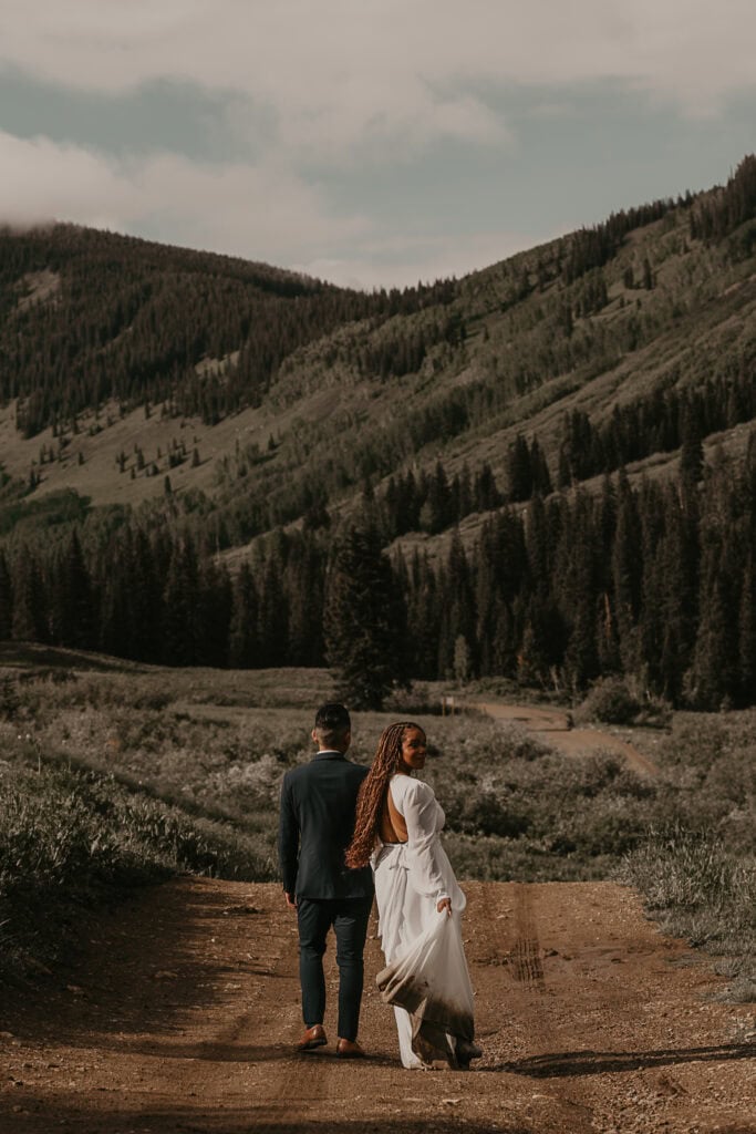 A couple in wedding attire walks towards mountains on a dirt path. The bride looks back at the camera.