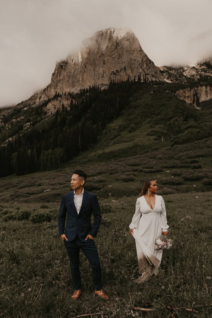 A couple stands in front of a fog covered mountain while looking opposite directions. They're dressed in wedding attire.