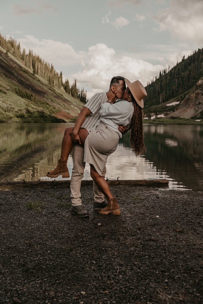 A man dip kisses a woman in front of an alpine lake.