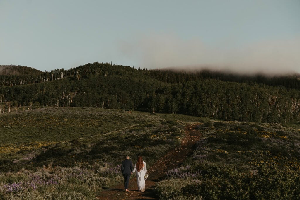 A bride and groom walk along a trail towards foggy mountains. Wildflowers surround them.