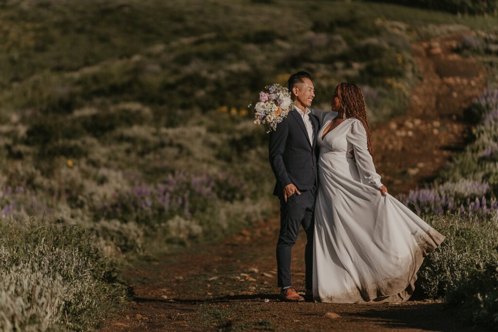 A bride and groom stand on a trail among wildflowers. She swings the muddy hem of her dress.