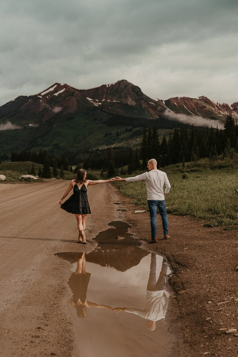 A couple reaches for each other while standing over a puddle that reflects them. Mountains are in the distance.