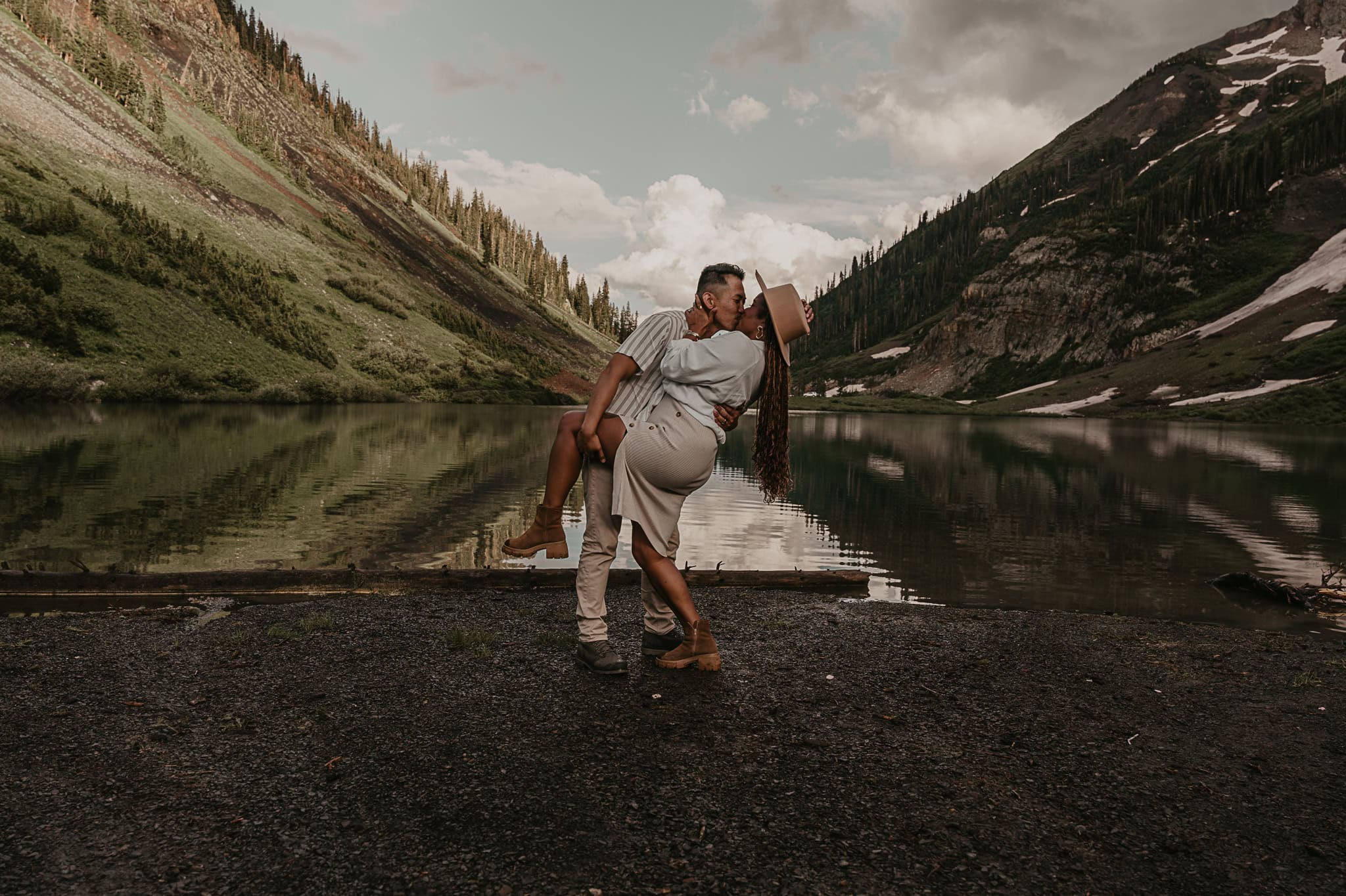 A couple kisses in front of an alpine lake that looks symmetrical as it reflects the mountains.